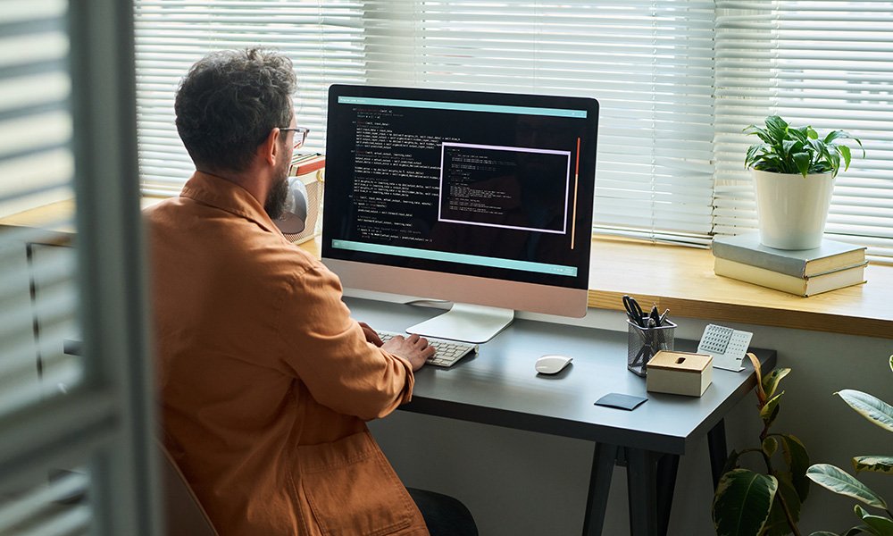 Man coding on a computer at a desk with blinds and plants